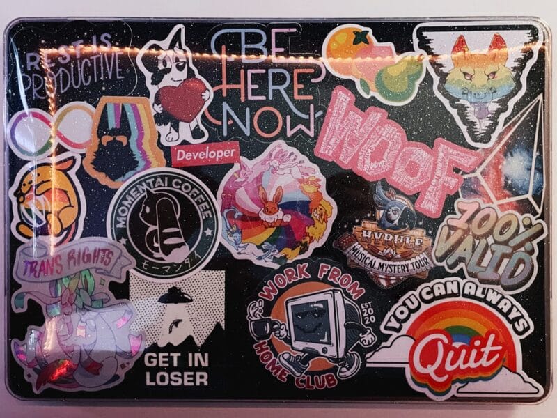 The back of a laptop with a sparkly plastic cover which is covered with a variety of stickers. The stickers are a mix of furry, pride, computer, and general nerdy stickers.